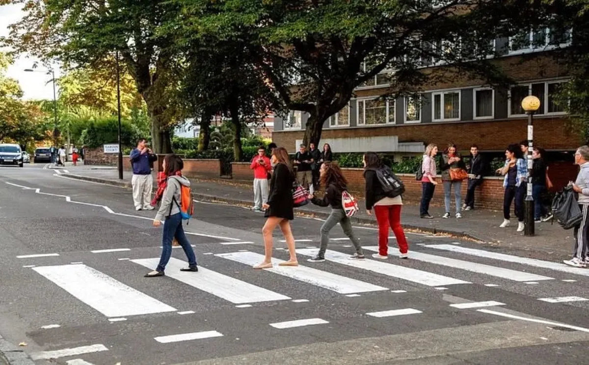 A group of people crossing the famous Abbey Road zebra crossing, with onlookers and surrounding trees visible.