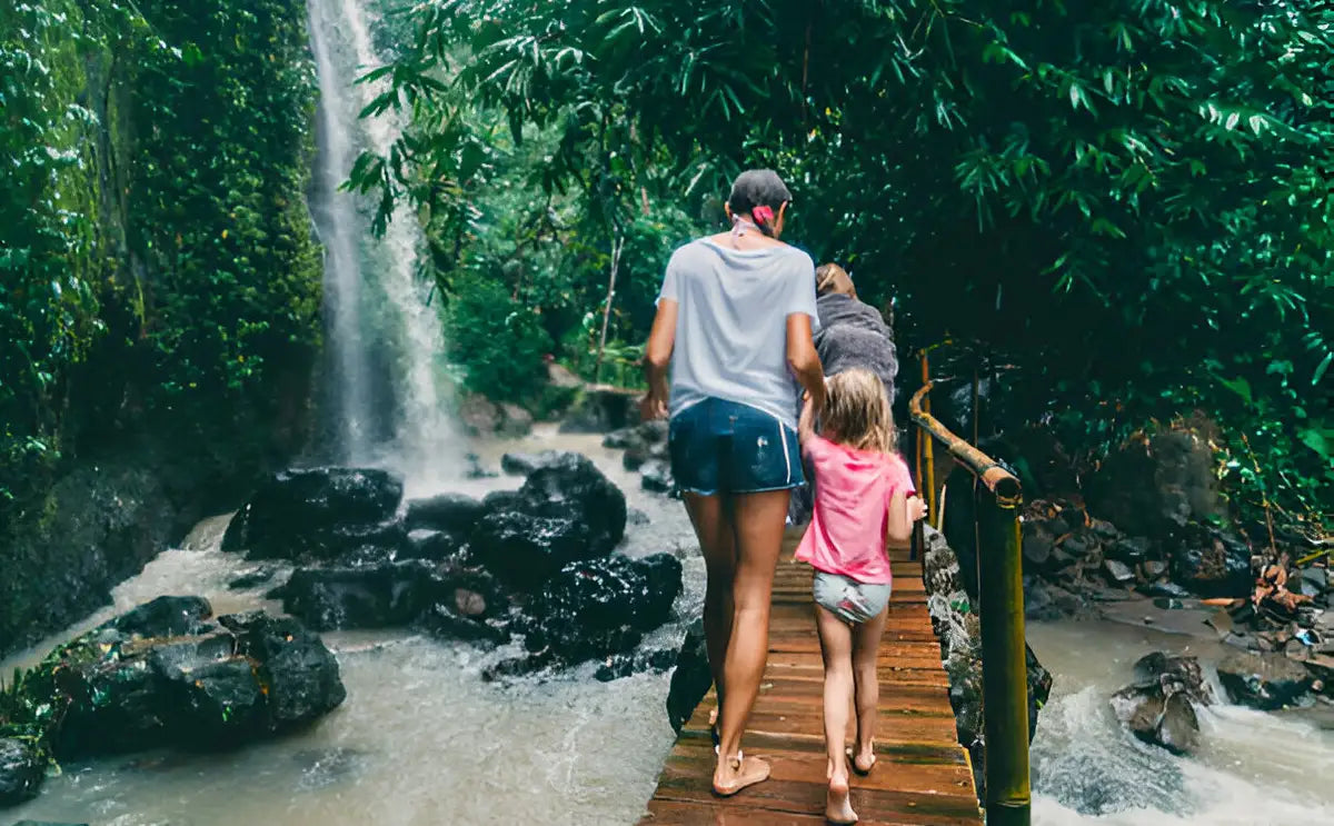A mother and her two children walk along a wooden bridge near a cascading waterfall surrounded by lush green foliage. The mist from the waterfall creates a refreshing atmosphere as they explore the tranquil natural setting. The scene captures the essence of a family adventure in the heart of Bali’s serene landscapes.