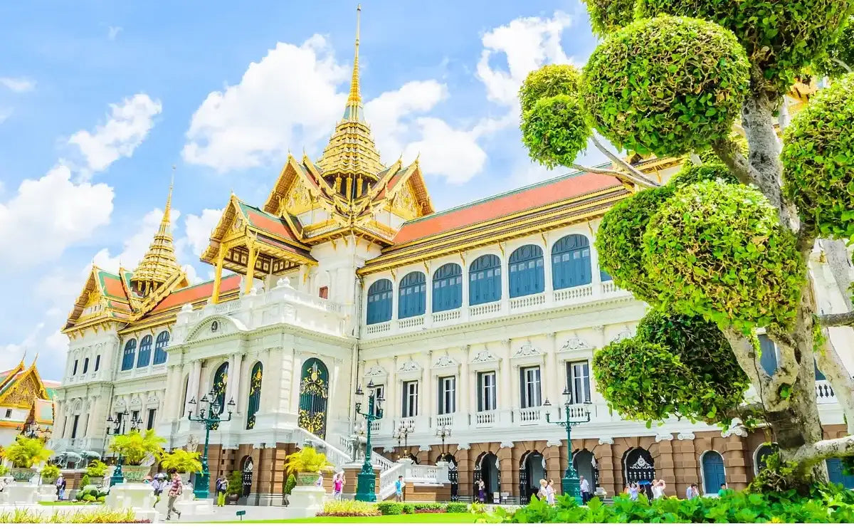 Exterior view of the Grand Palace in Bangkok, showcasing its ornate architecture, golden rooftops, and manicured trees under a bright blue sky.