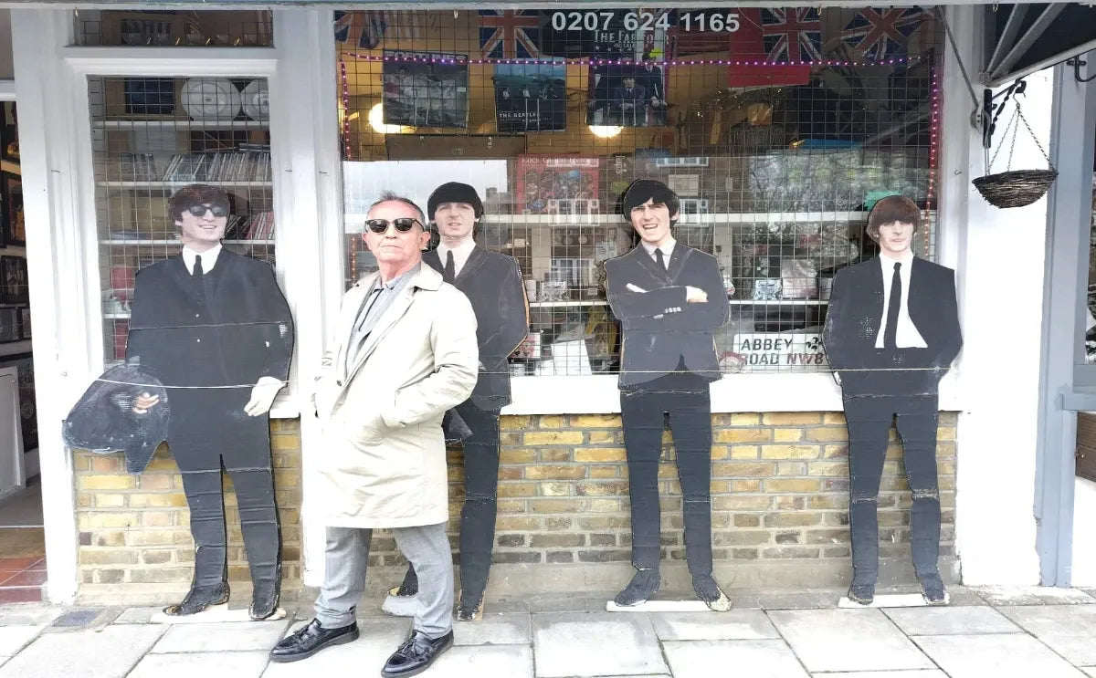 A man posing confidently outside a store with life-sized cutouts of The Beatles members in suits displayed in the background.