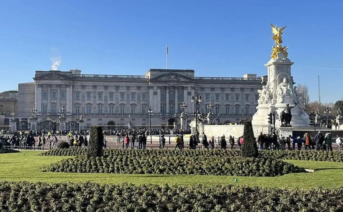 Buckingham Palace on a sunny day, with the Victoria Memorial in the foreground surrounded by neatly trimmed gardens and visitors exploring the area.