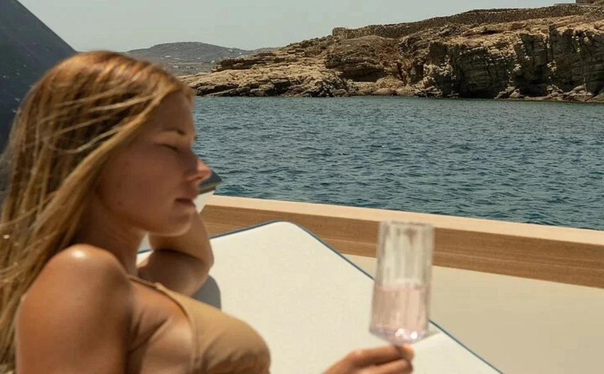 A woman relaxes on the deck of a "Nevma" luxury yacht in Mykonos, enjoying a glass of champagne as the scenic rocky coastline stretches in the background. The tranquil moment captures the essence of a private, serene getaway.