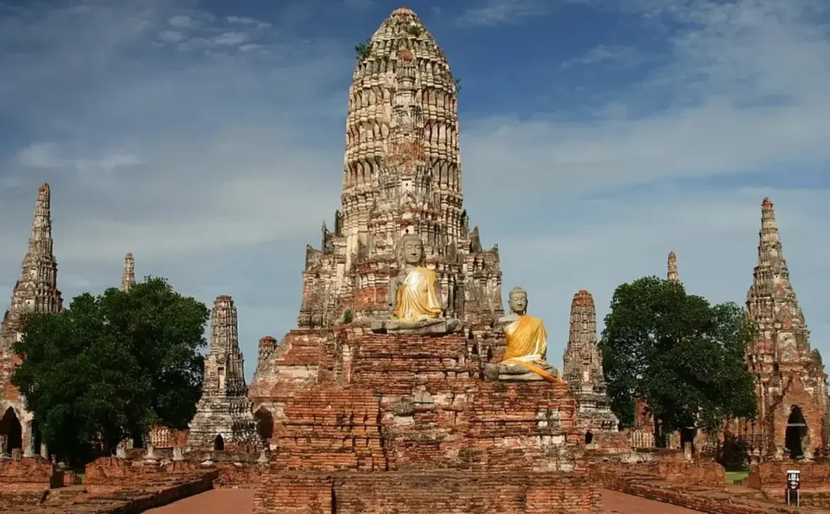 Front view of Wat Chaiwatthanaram in Ayutthaya Historical Park, featuring two large Buddha statues draped in yellow robes seated in front of the central prang.