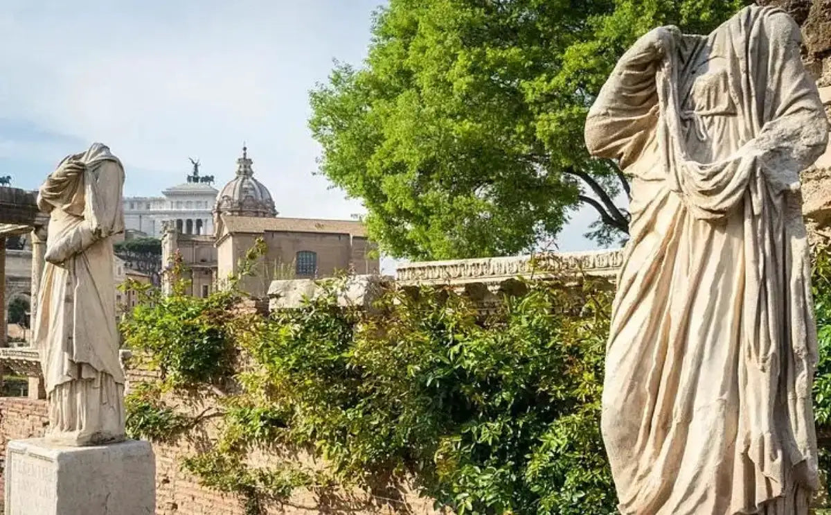 Close-up of two ancient headless statues in the Roman Forum, surrounded by greenery and historic stone walls. In the background, a large dome and the Vittorio Emanuele II Monument are visible under a clear sky.
