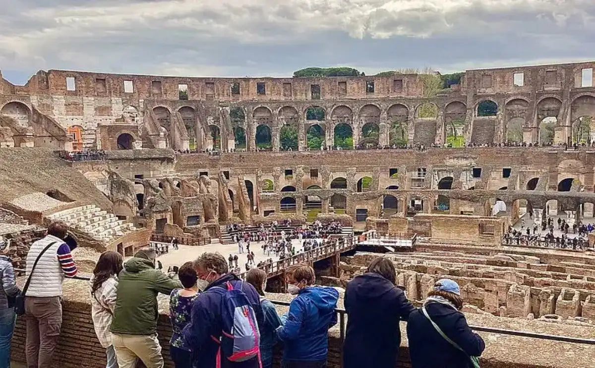 Interior view of the Colosseum in Rome, showing the ancient stone walls, arches, and reconstructed wooden platforms. Visitors stand along the viewing balcony in the foreground, overlooking the central arena and lower levels. Overcast sky in the background.
