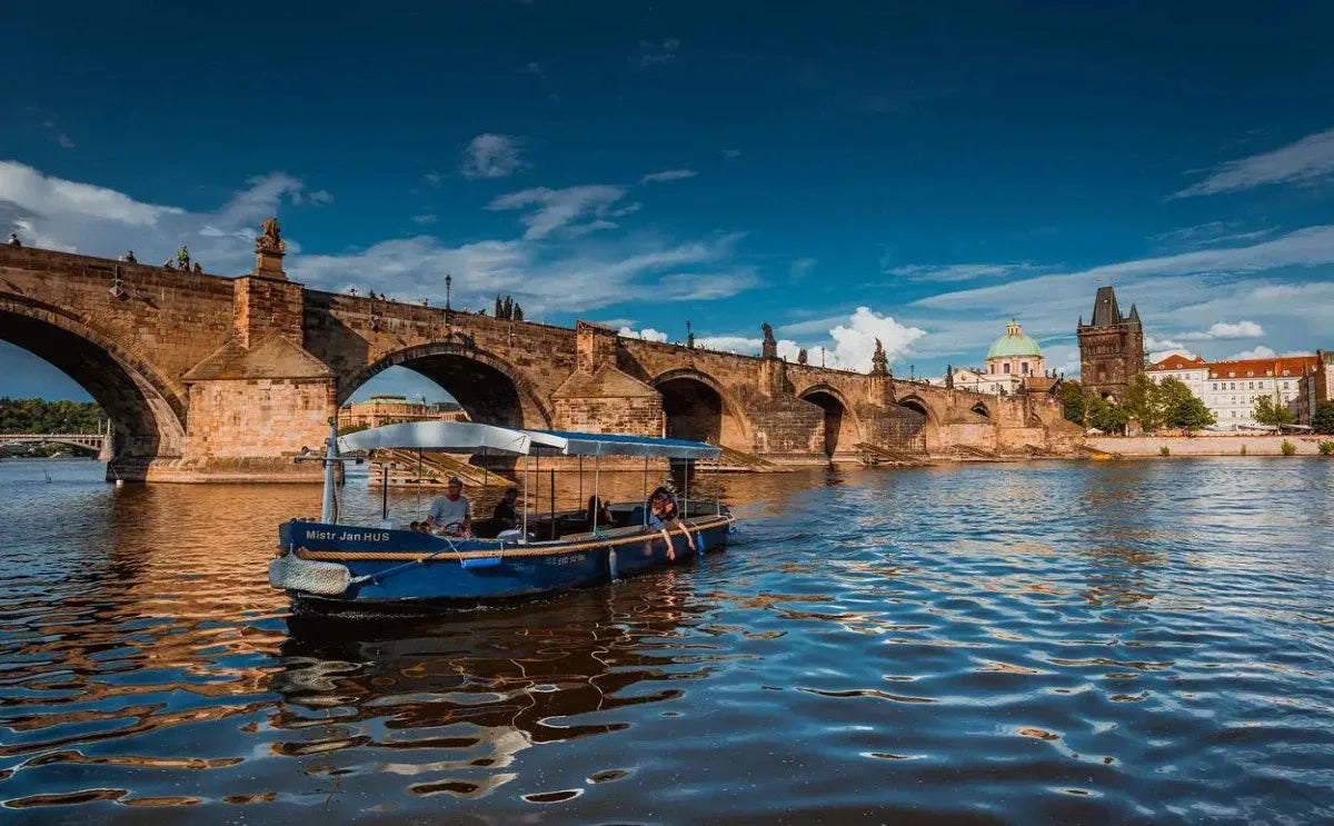 A small blue boat named "Mistr Jan Hus" sails on the Vltava River near the historic Charles Bridge in Prague. The iconic bridge's stone arches stretch across the river, with the Old Town Bridge Tower and dome of St. Francis of Assisi Church visible in the background under a bright blue sky.
