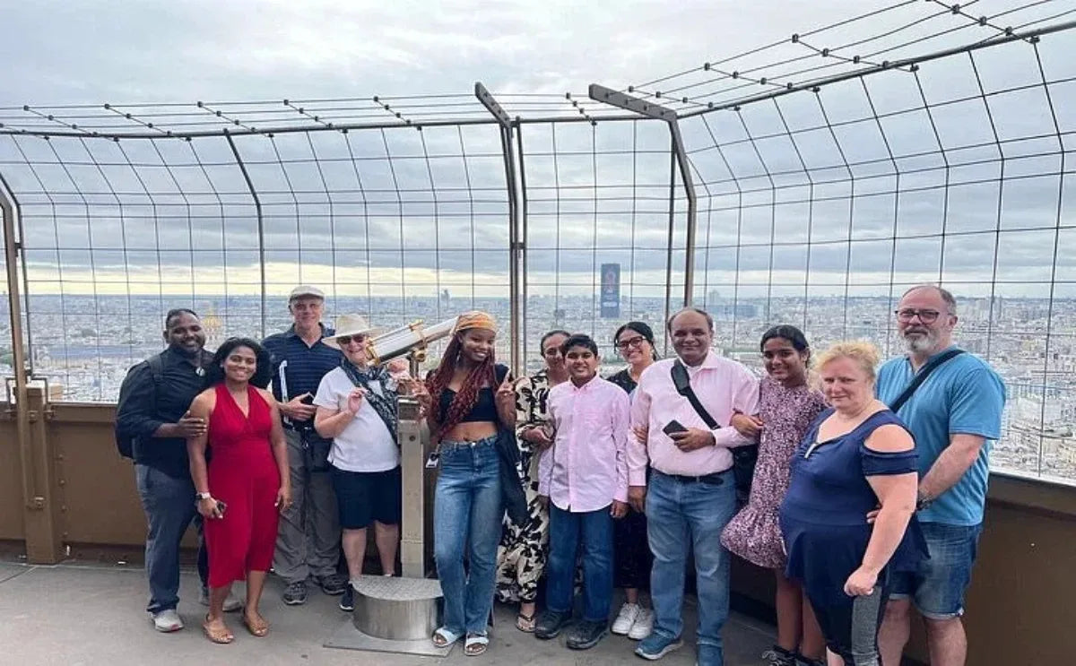 Group of visitors posing for a photo at the Eiffel Tower observation deck, with panoramic views of Paris in the background.