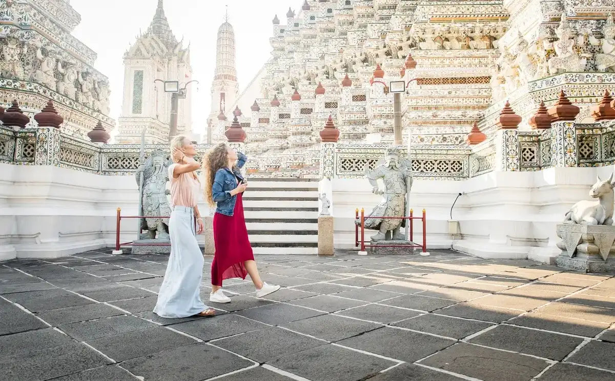 Two female tourists walking and pointing at the intricate details of the Wat Arun temple complex during the Grand Palace and temples tour in Bangkok.