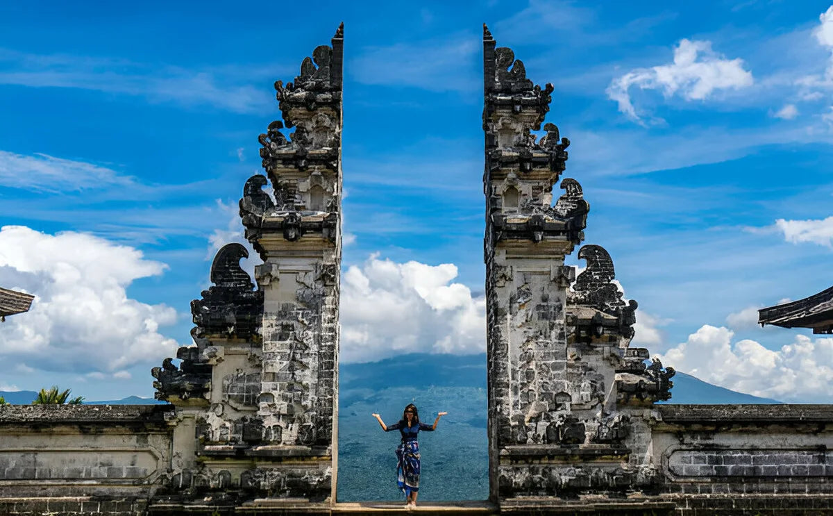 A person stands between the iconic split gates of Lempuyang Temple in Bali, with a backdrop of clear blue skies and distant mountains, symbolizing the 'Gates of Heaven.