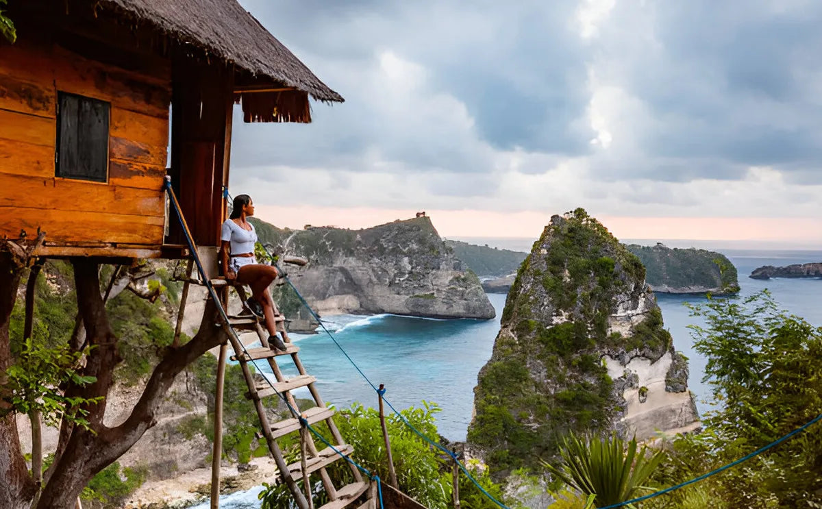 Brunette woman sitting on the steps of a wooden treehouse overlooking the stunning cliffs and turquoise waters of Nusa Penida, Bali, during sunrise.