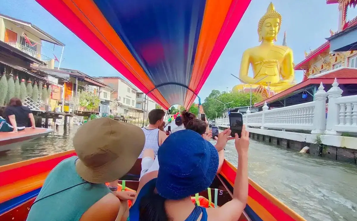 Tourists riding in a colorful longtail boat along a canal in Bangkok, capturing photos of the golden Buddha statue and traditional Thai architecture in the background, as they pass by stilted houses lining the waterway.