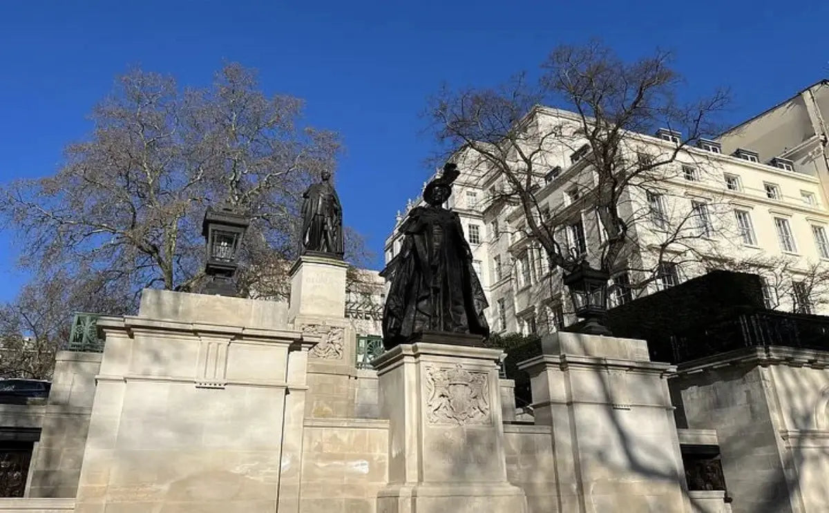 Statues of historical figures in a formal setting, framed by a clear blue sky, bare trees, and classic architectural buildings in the background.