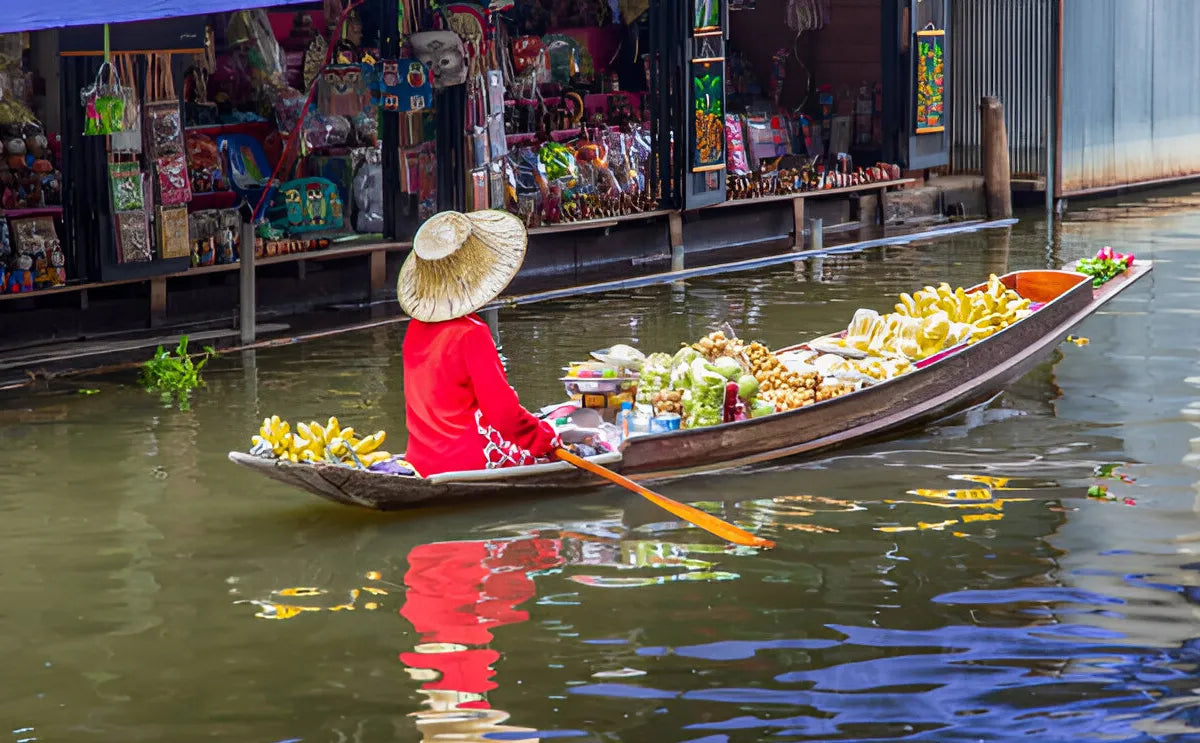 An elderly Asian woman wearing a hat, selling a variety of colorful Thai fruits from her boat at the bustling Damnoen Saduak Floating Market in Ratchaburi.