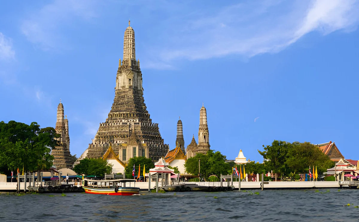 Scenic view of Wat Arun, the Temple of Dawn, set against a bright blue sky, with a colorful boat floating on the Chao Phraya River, enhancing the vibrant atmosphere of Bangkok, Thailand.