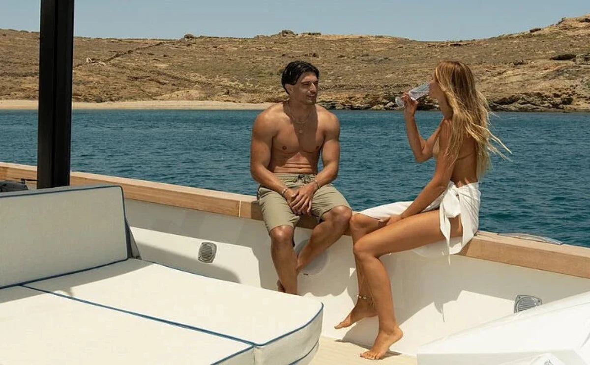A man and a woman enjoy a relaxed moment on a "Nevma" boat with crystal-clear waters and a sandy coastline in the background. The serene ambiance highlights the allure of a private cruise in Mykonos.