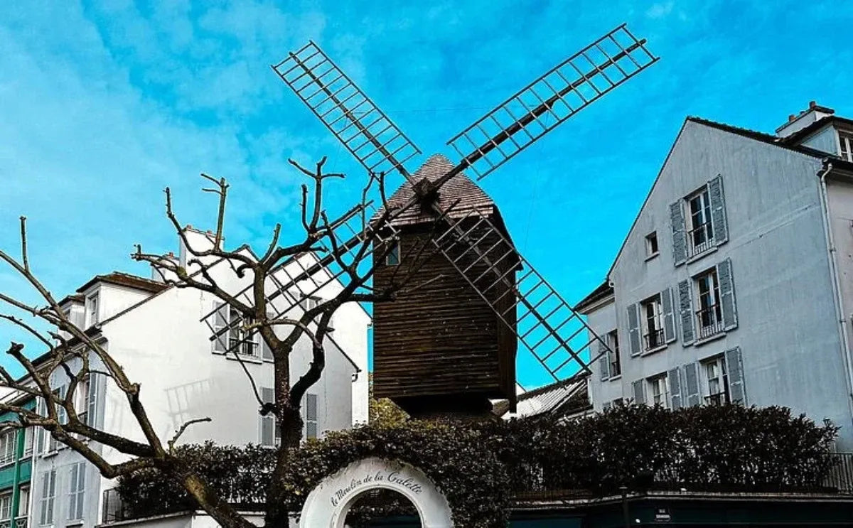 Historic windmill surrounded by trees and residential buildings in Montmartre, with a clear blue sky above.