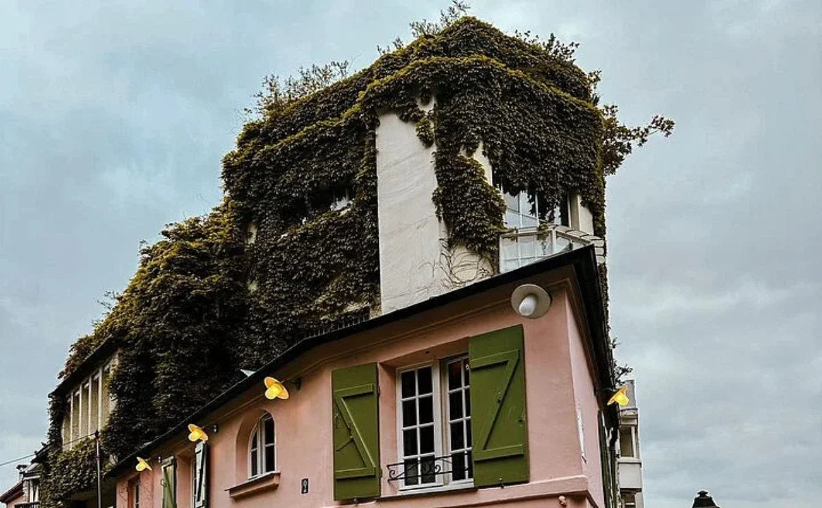 Building corner in Montmartre with ivy-covered walls and a pink lower facade featuring green shutters under a cloudy sky.
