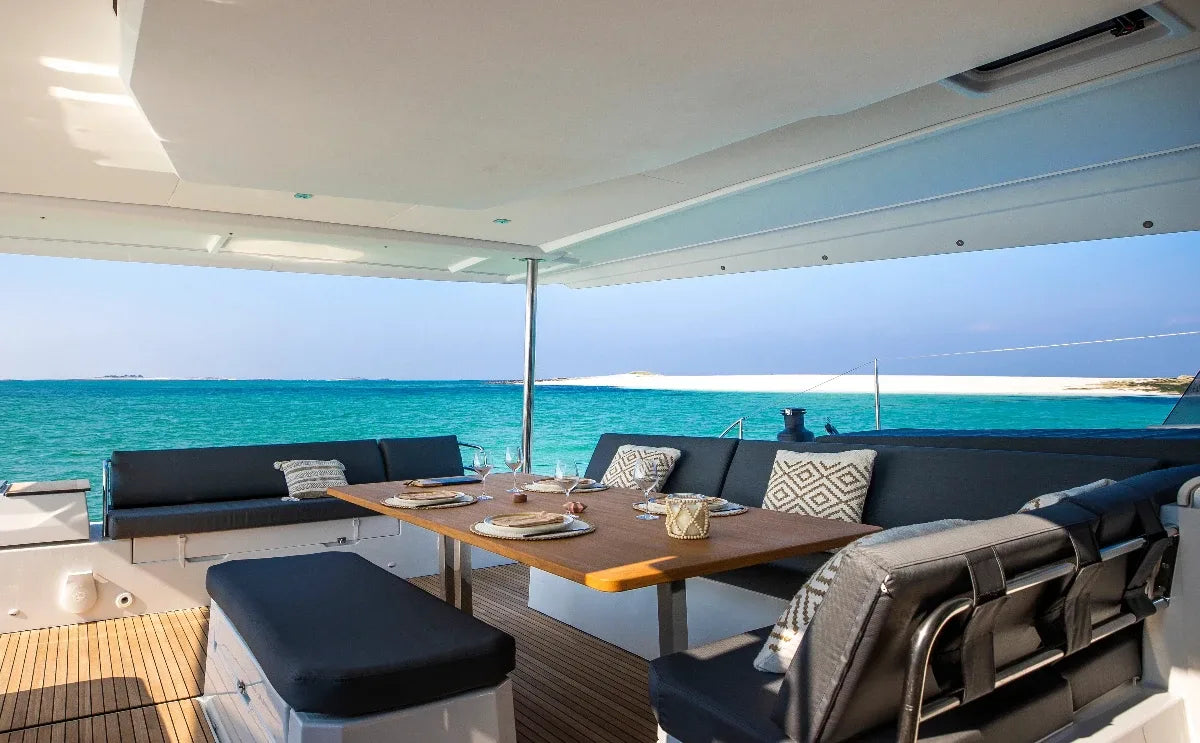 A cozy dining area on a sailing catamaran with a wooden table set for a meal, overlooking crystal-clear turquoise waters and a sandy shoreline in the distance.