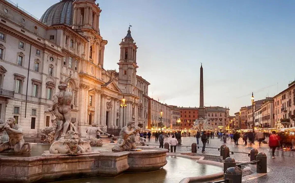 Piazza Navona at dusk in Rome, featuring the ornate Fountain of Neptune in the foreground and the Baroque church of Sant'Agnese in Agone illuminated by soft lights. The square is lively with people strolling and dining at nearby cafes, with the Egyptian obelisk standing prominently in the center.