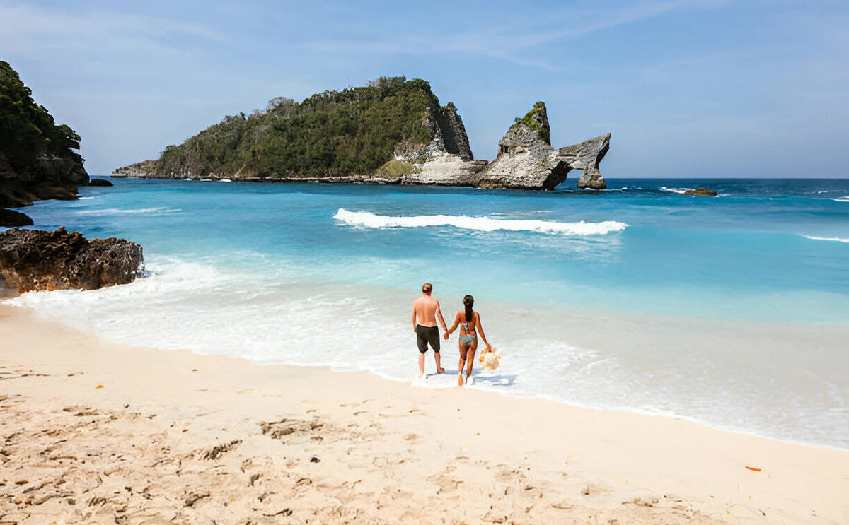 Couple walking hand in hand along the pristine sands of Atuh Beach in Nusa Penida, Bali, with clear turquoise waters and dramatic rock formations in the background.