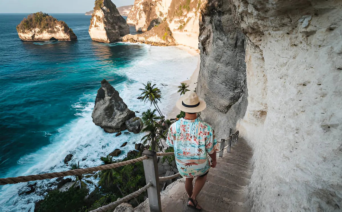 Male tourist with flower pattern shirt walking down the carved staircase to Diamond Beach, surrounded by dramatic cliffs, turquoise waters, and palm trees in Nusa Penida, Bali.