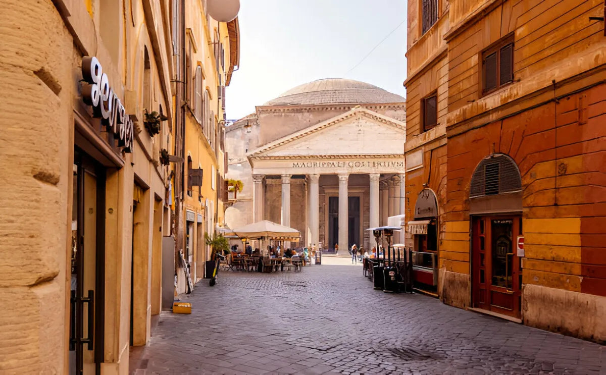 View of the Pantheon in Rome from a narrow cobblestone street lined with colorful buildings. The iconic temple with its grand columns and triangular pediment dominates the scene, with outdoor café tables and pedestrians visible near the entrance under a bright afternoon sky.