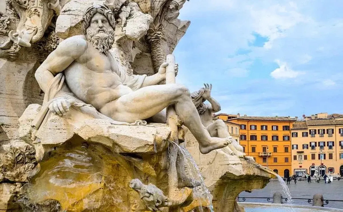 Close-up of a sculpted figure from the Fountain of the Four Rivers in Piazza Navona, Rome. The detailed statue represents a reclining river god, holding a paddle, with water cascading into the basin below. In the background, colorful Roman buildings line the square under a bright, partly cloudy sky.