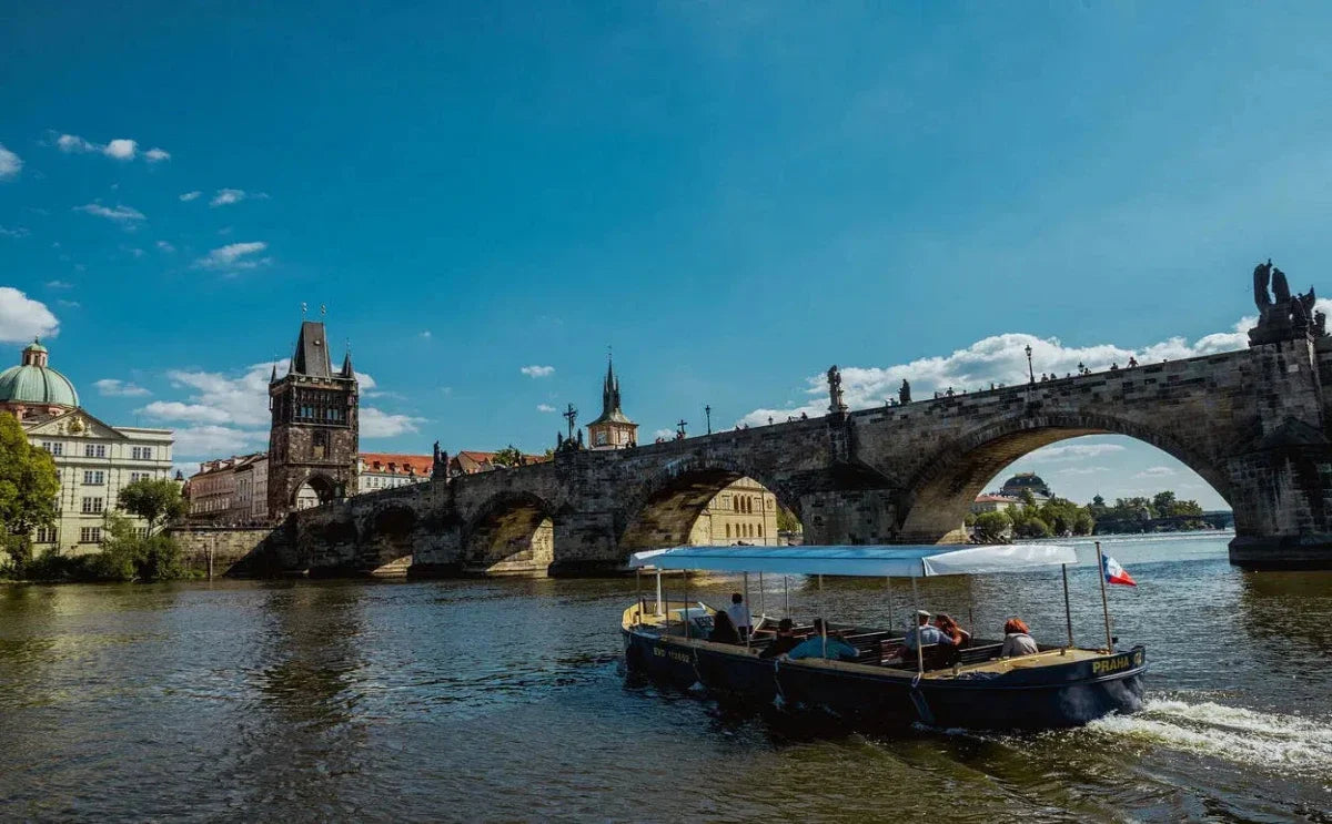 A small boat with a Czech flag sails on the Vltava River near the iconic Charles Bridge in Prague. The bridge's historic stone arches are complemented by the Old Town Bridge Tower and surrounding Gothic and Baroque architecture under a clear blue sky.