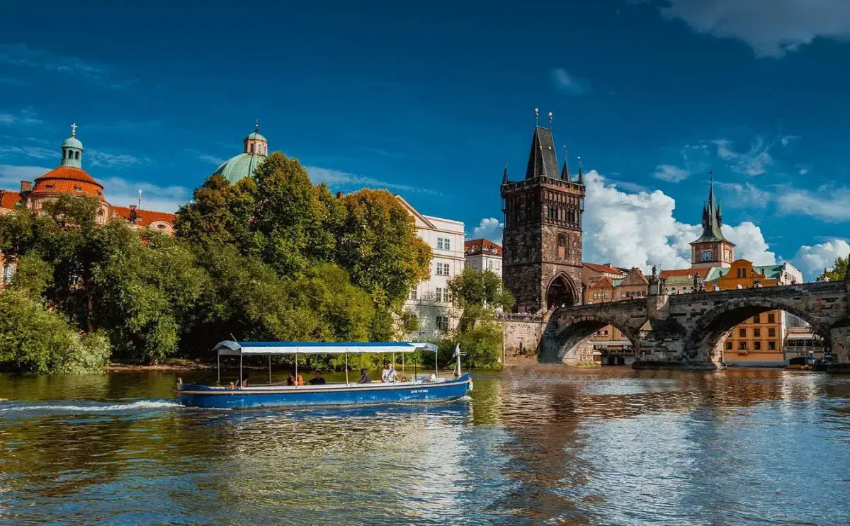 A blue boat cruises along the Vltava River, passing near the Charles Bridge and the Old Town Bridge Tower in Prague. The scenic view includes lush greenery, domed rooftops, and Gothic architecture, all under a vivid blue sky with scattered clouds.