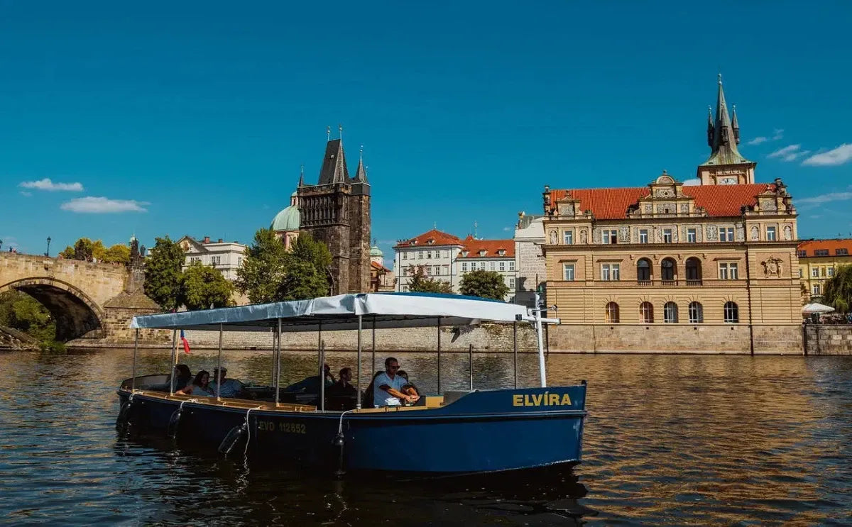A small blue boat named "Elvíra" floats on the Vltava River near Prague's historic Old Town. In the background, the Charles Bridge's stone arch and the Gothic tower of the Old Town Bridge Tower rise alongside a Baroque-style building with a red-tiled roof under a clear blue sky.