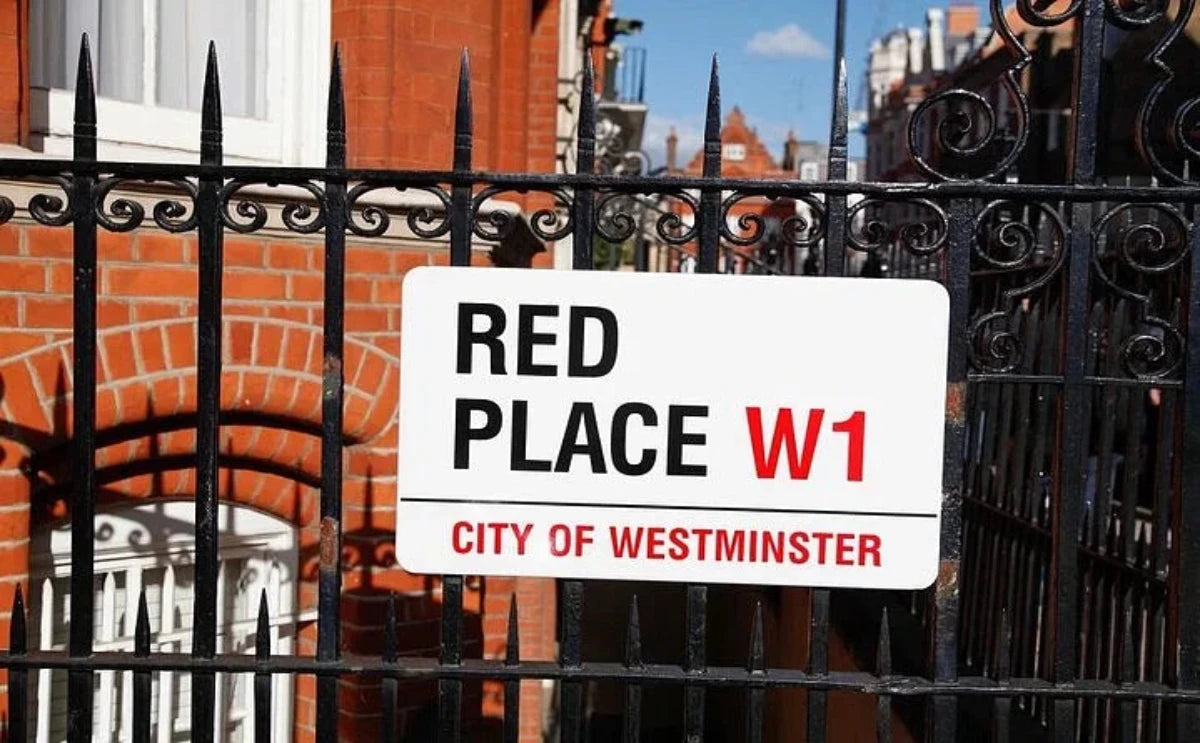 A street sign reading "Red Place W1 City of Westminster," mounted on a black wrought-iron fence with a red-brick building in the background.