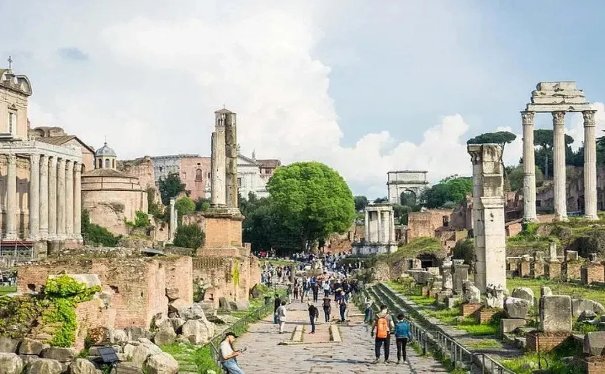 Ground-level view of the Roman Forum in Rome, showcasing ruins including columns, scattered stones, and ancient walls. Visitors walk along a central pathway, surrounded by historical structures and greenery under a partly cloudy sky.