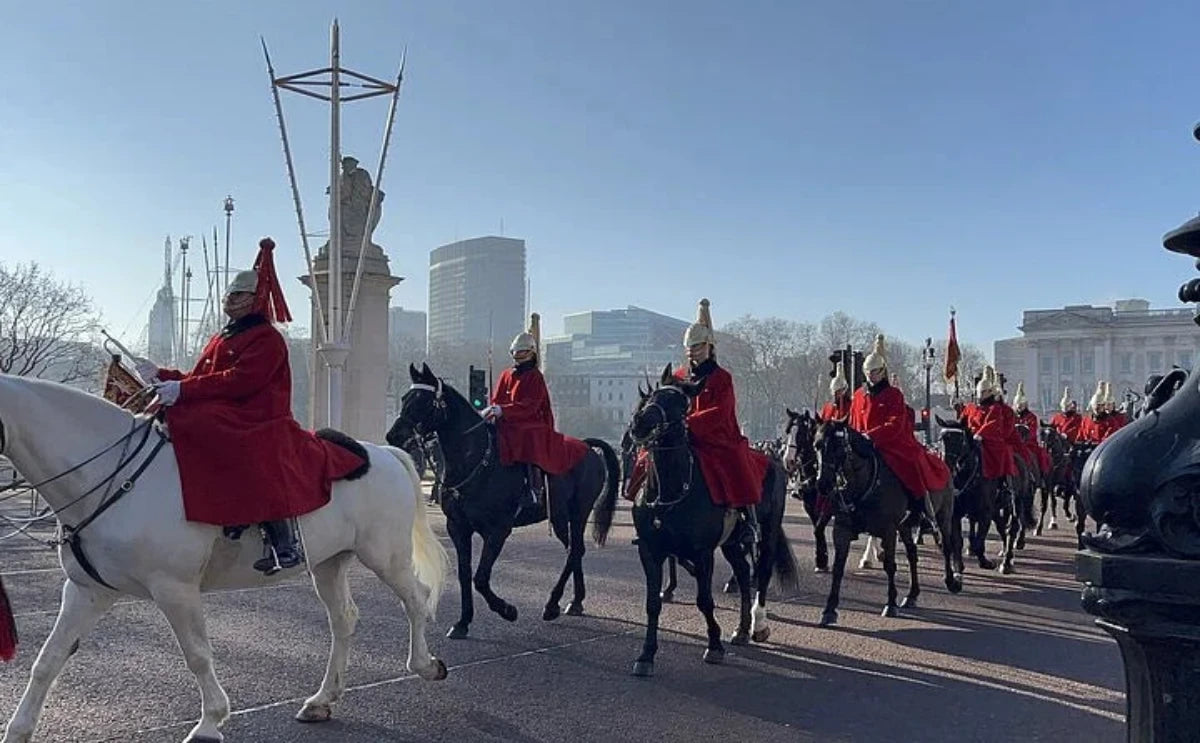 A procession of mounted royal guards dressed in red coats and helmets, riding horses near a landmark in London, with city buildings in the background.