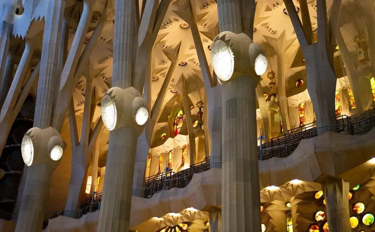 Interior view of the Sagrada Familia in Barcelona, showing the intricate columns resembling trees, illuminated with warm and colorful light from the stained glass windows.