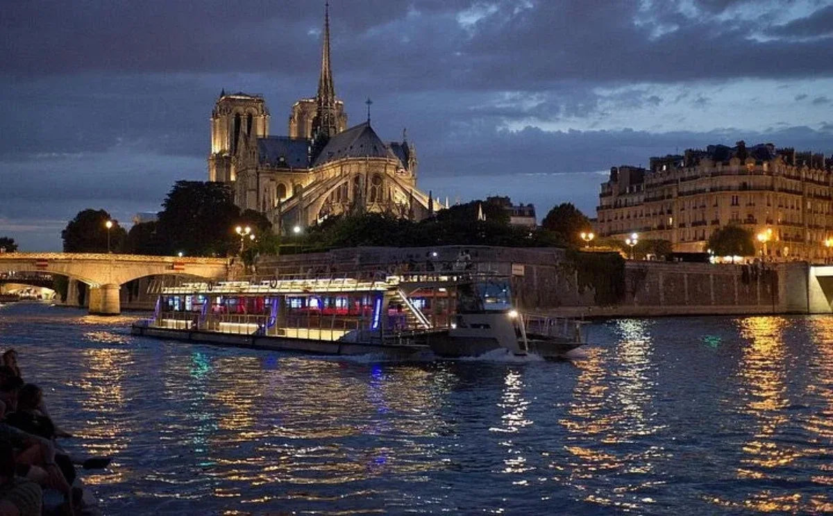 A Seine river boat illuminated with colorful lights cruising at night near the Notre Dame Cathedral, with reflections shimmering on the water and Parisian buildings in the background.