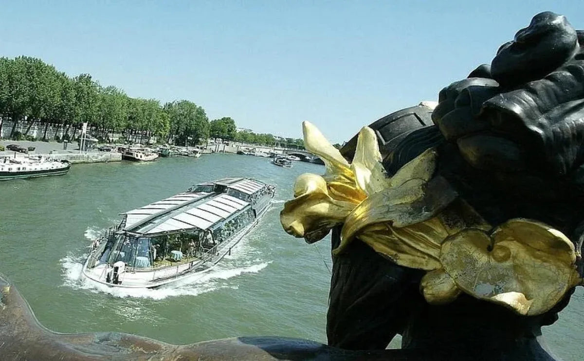 A sightseeing boat cruising along the Seine River in Paris, surrounded by green riverbanks and moored boats, viewed from the Pont Alexandre III bridge with part of its ornate golden sculpture in the foreground.