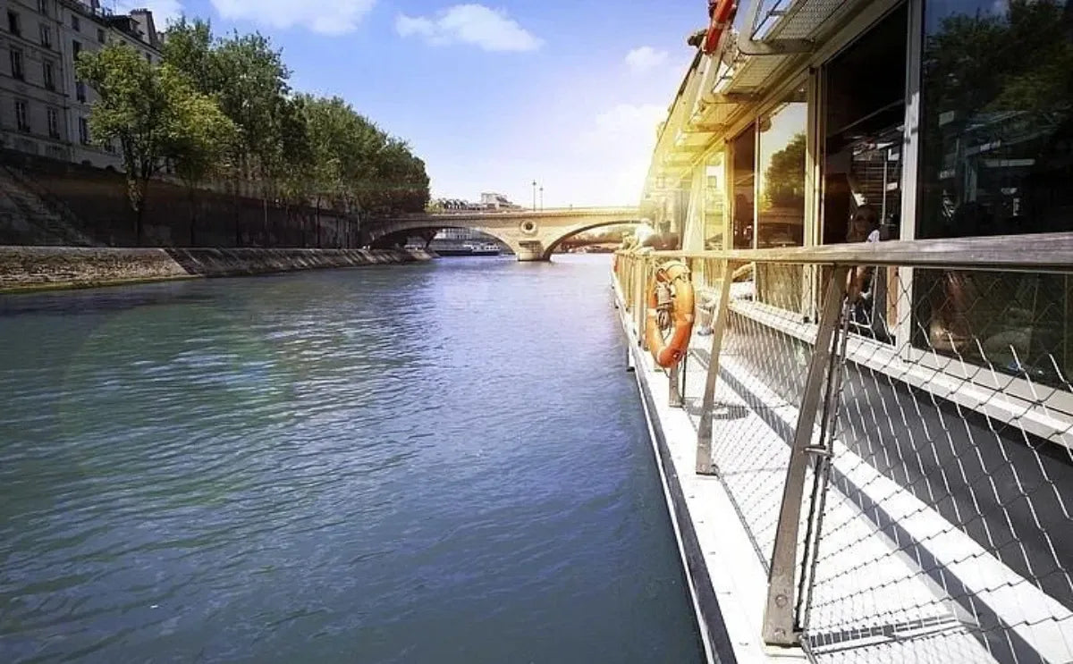 Side view of a Seine River cruise boat in Paris, with sunlight reflecting off the water and a nearby bridge in the distance, framed by trees and riverside buildings.