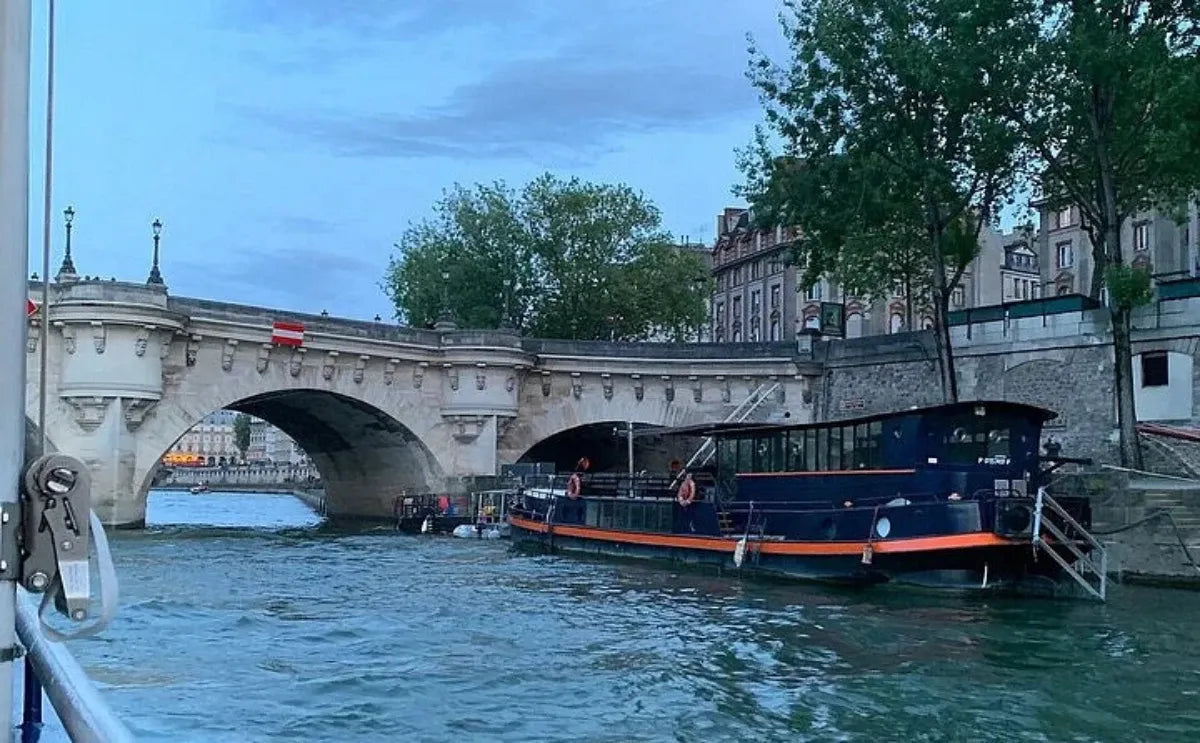A docked riverboat on the Seine River in Paris near the Pont Neuf bridge, with stone arches and nearby trees lining the riverbank under a cloudy evening sky.