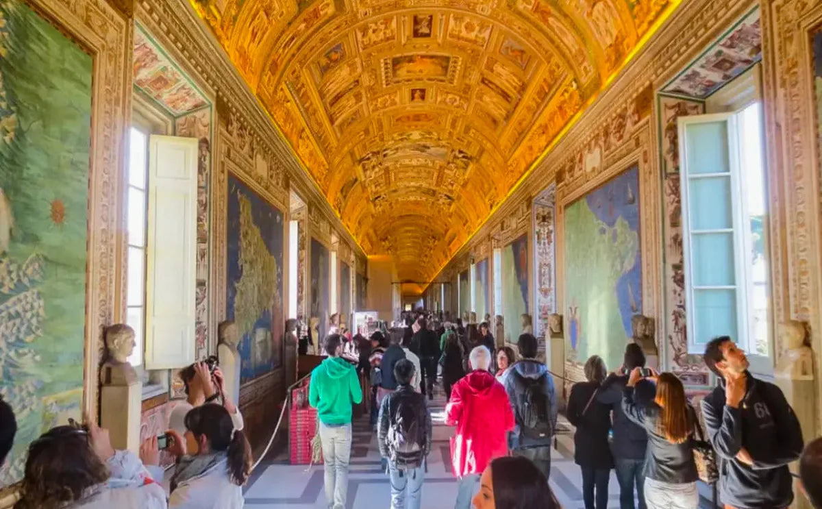 Visitors exploring the Gallery of Maps in the Vatican Museums, showcasing detailed frescoed maps on the walls and a richly decorated arched ceiling illuminated with warm lighting.