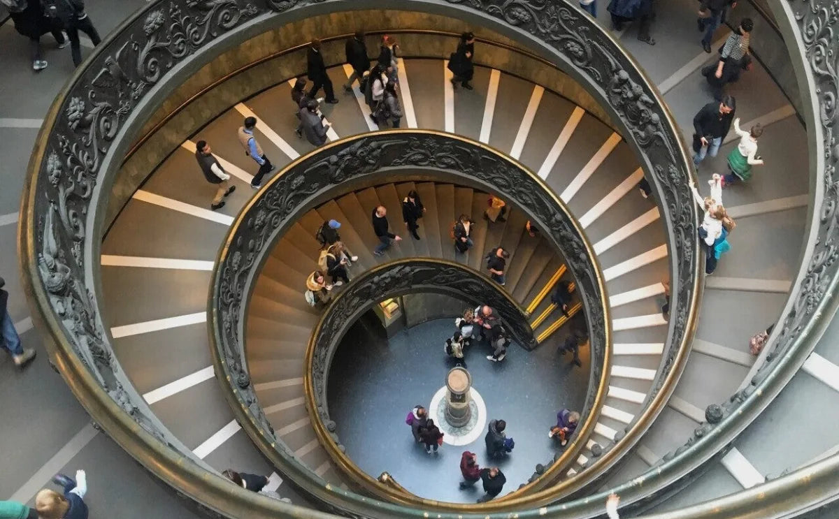 Spiral staircase at the Vatican Museums, featuring ornate railings and a symmetrical design, with visitors descending and ascending the steps. The view is captured from above, showcasing the intricate architecture.