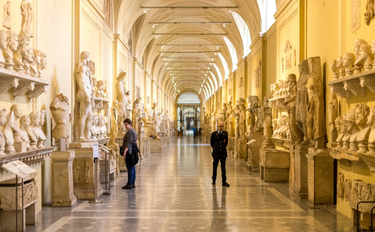 Interior of the Vatican Museums with rows of ancient sculptures and busts displayed along both sides of a long, arched corridor illuminated by warm lighting, featuring visitors observing the exhibits.