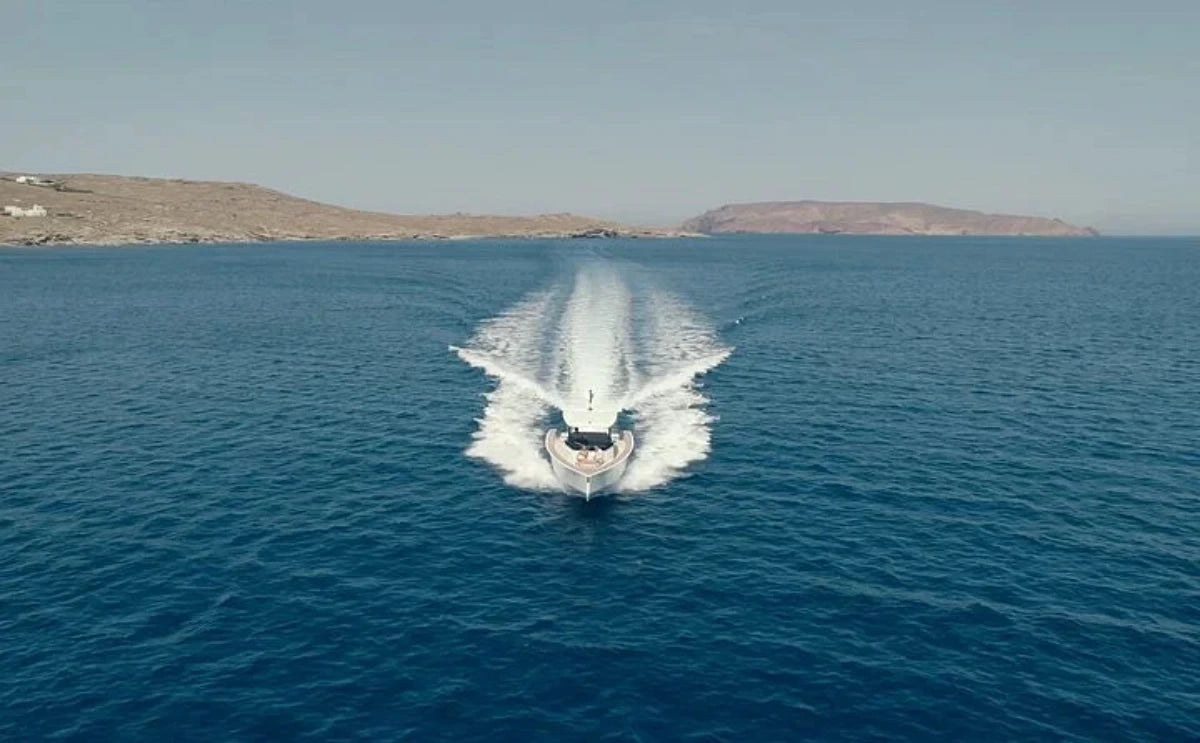 "Nevma" speeds across the clear blue waters of Mykonos, leaving a foamy trail behind. The scenic coastline with rugged hills can be seen in the distance under the bright, sunny sky.