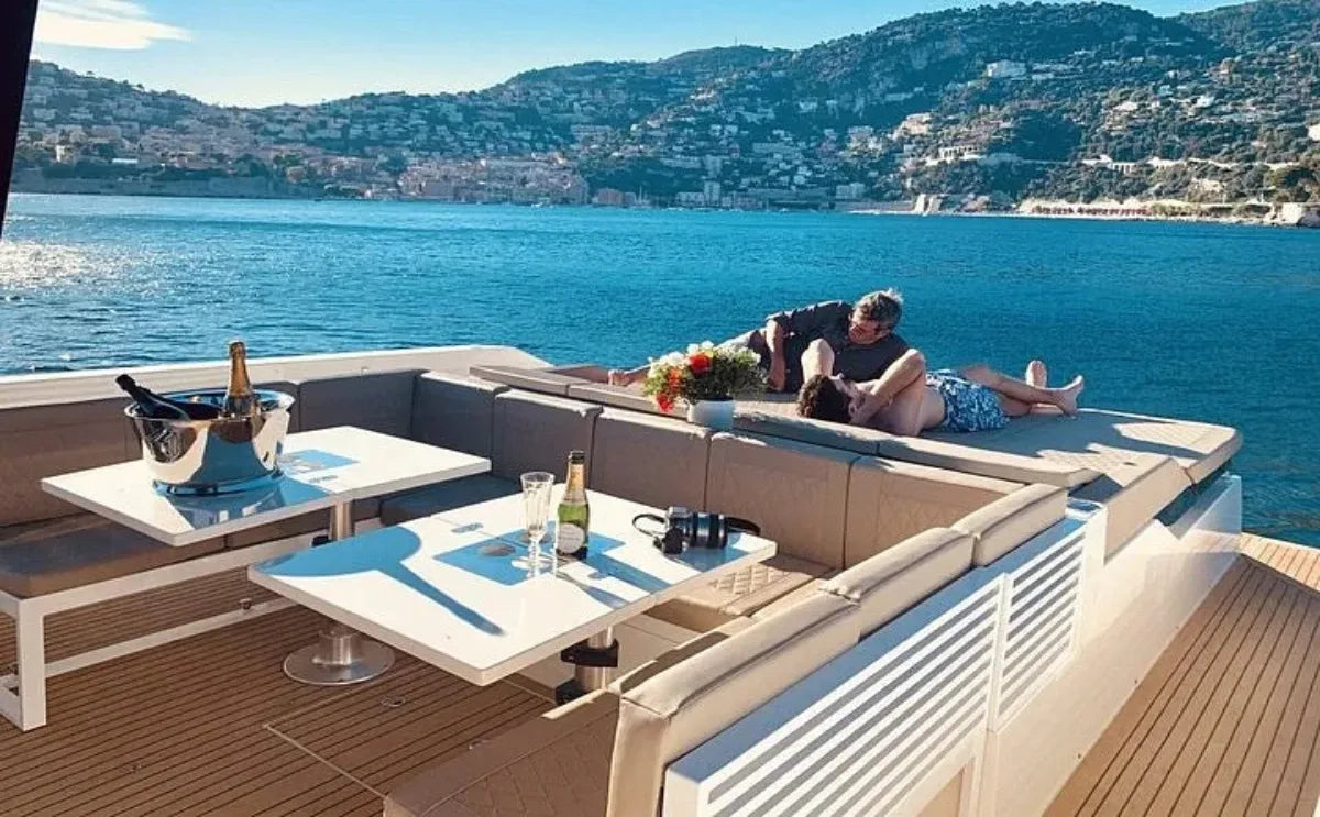 A luxurious yacht deck featuring comfortable seating and a table with champagne and glasses, set against a scenic coastal backdrop. A couple relaxes on the sunbed, enjoying the serene views of Mykonos in the distance.