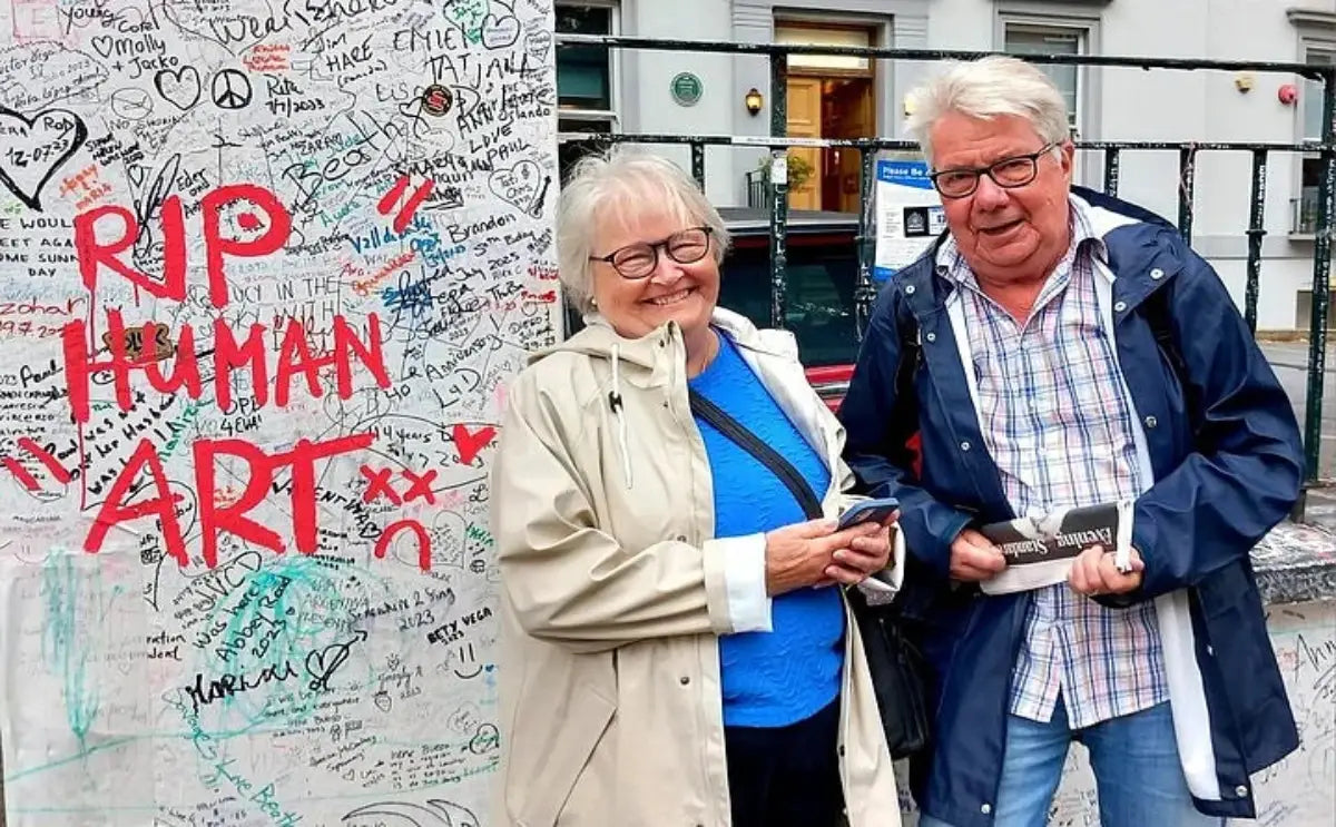 Two Beatles fans standing near a graffiti-covered wall, smiling, and holding memorabilia, likely near Abbey Road Studios.