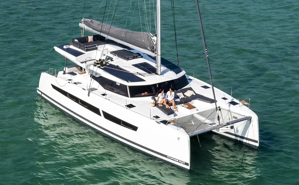 A modern sailing catamaran floats on serene turquoise waters, with two passengers relaxing on its spacious deck, enjoying the open sea views and sunlight.