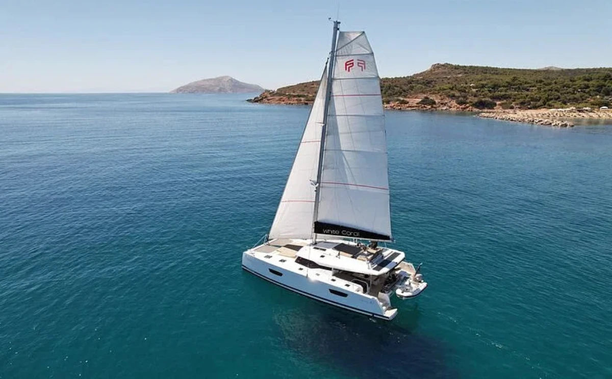 A sleek sailing catamaran named "White Coral" glides across calm blue waters, with a rocky coastline and distant hills framing the horizon under a clear sky.