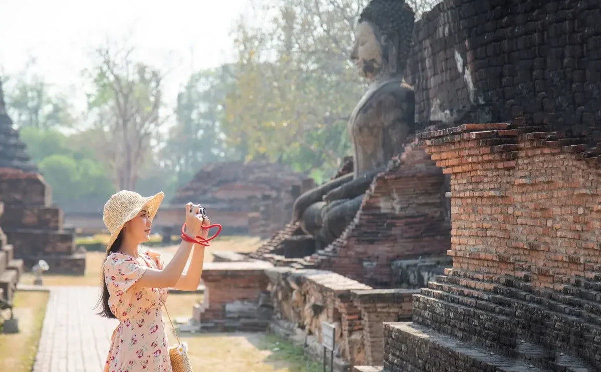 Female asian tourist photographing an ancient seated Buddha statue at Ayutthaya Historical Park, surrounded by weathered brick ruins and greenery.