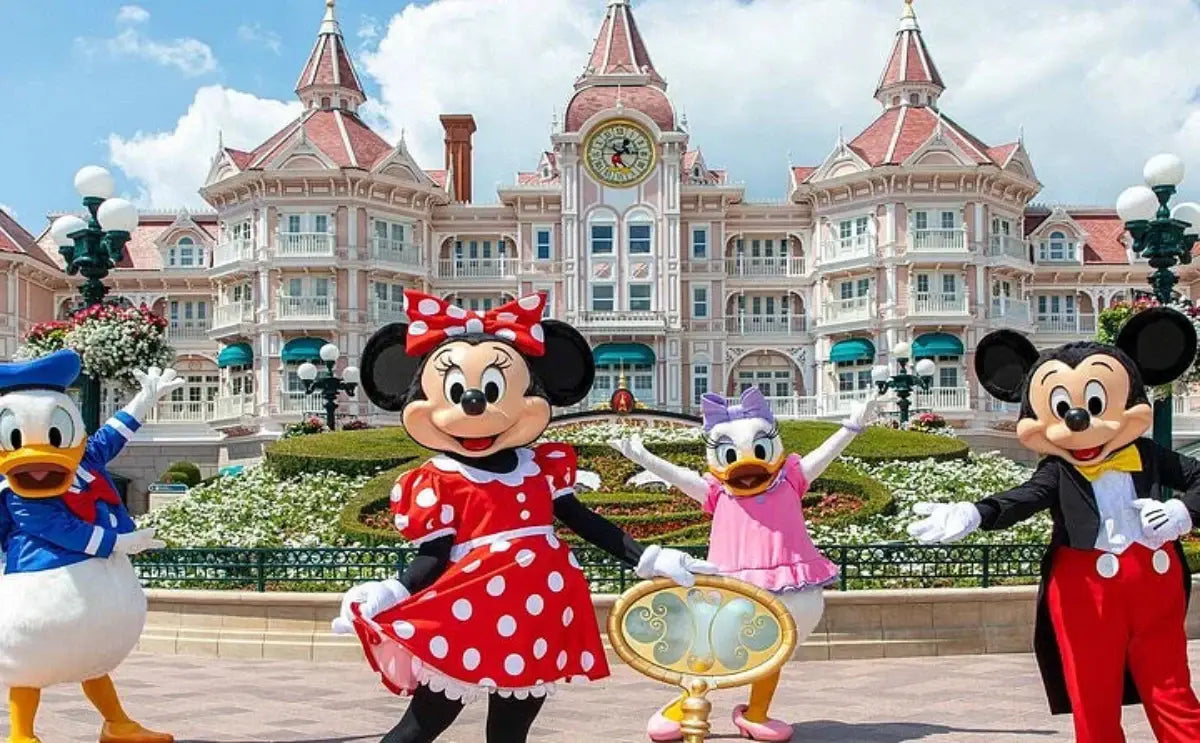 A vibrant scene at Disneyland Paris featuring iconic characters Mickey Mouse, Minnie Mouse, Donald Duck, and Daisy Duck posing in front of the enchanting pink Victorian-style Disneyland Hotel, surrounded by colorful flowers and cheerful ambiance.