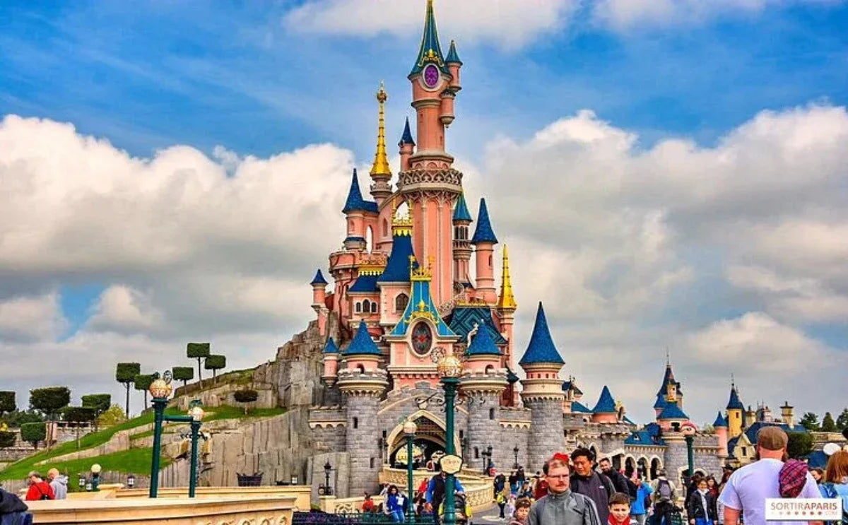 The magnificent Sleeping Beauty Castle in Disneyland Paris, with its iconic pink and blue spires, stands tall under a partly cloudy sky, surrounded by excited visitors exploring the magical surroundings.