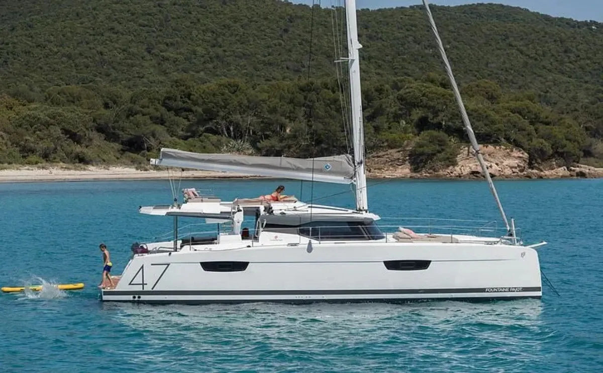 A modern sailing catamaran anchored in turquoise waters near a lush, green coastline. A child enjoys the water from a floating mat while an adult relaxes on the upper deck, soaking in the sun.